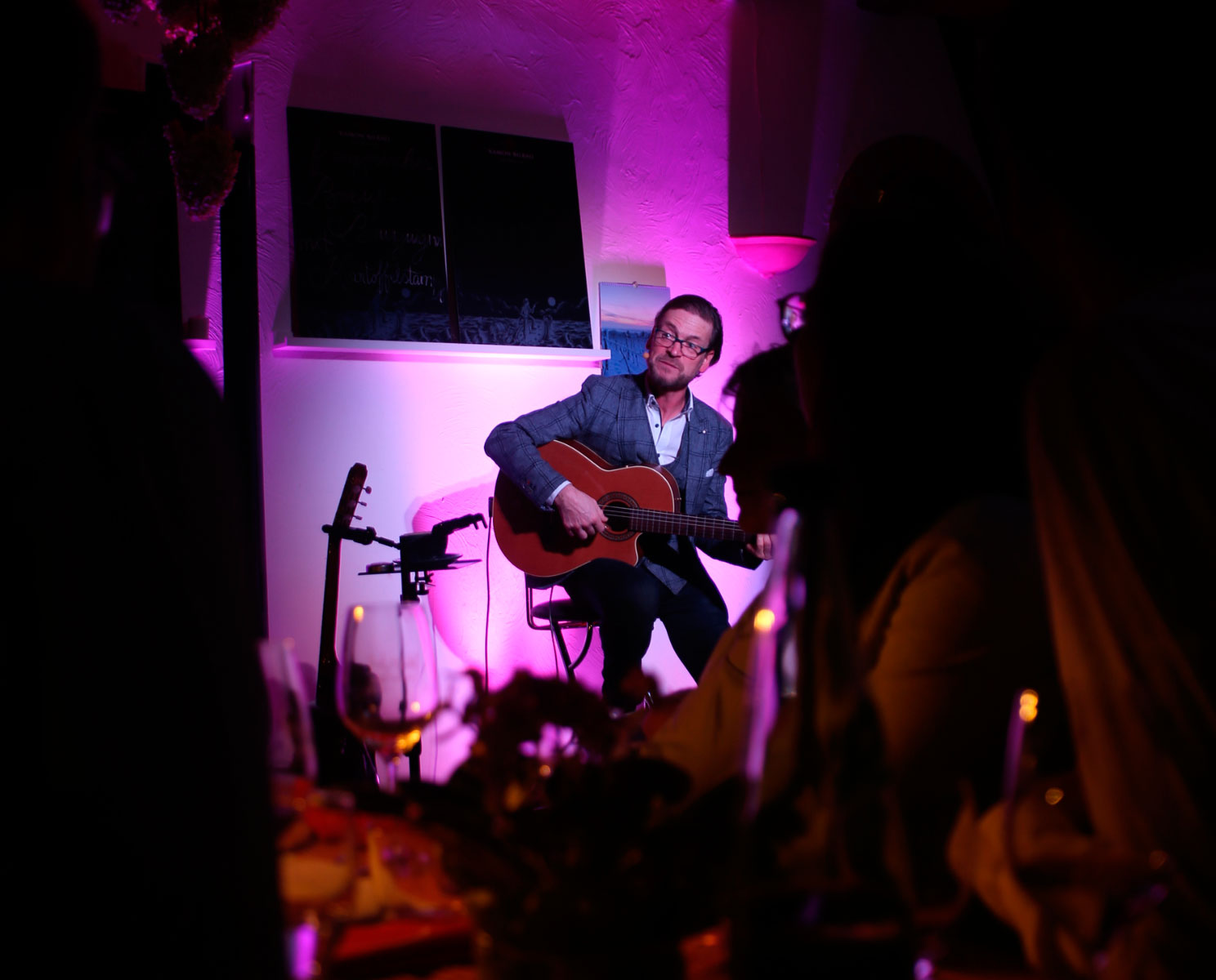 Mann sitzt auf einem Hocker und spielt Gitarre im Vordergrund des Bildes sitzen Menschen und schauen ihn an. 