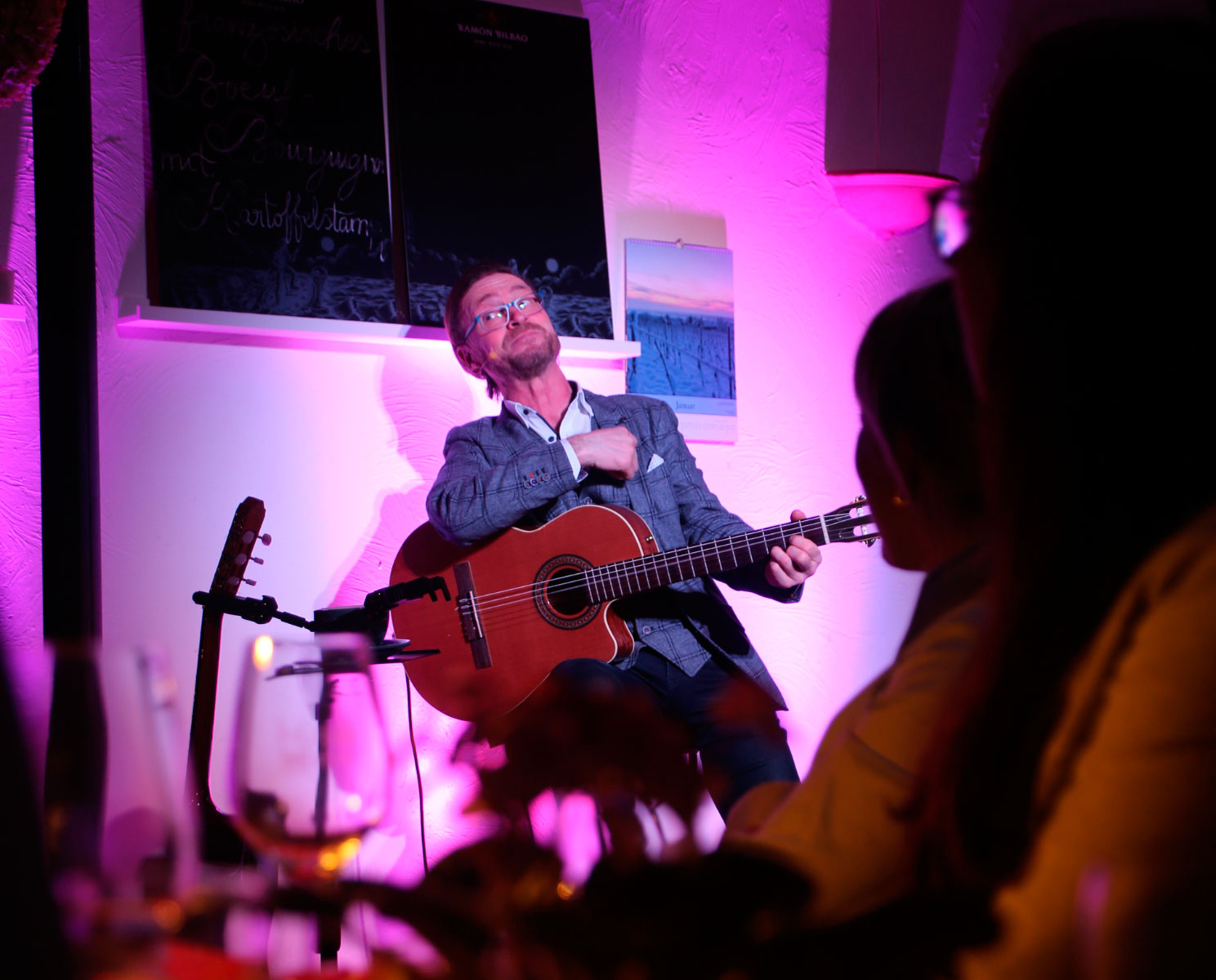 Mann sitzt auf einem Hocker und spielt Gitarre im Vordergrund des Bildes sitzen Menschen und schauen ihn an. 