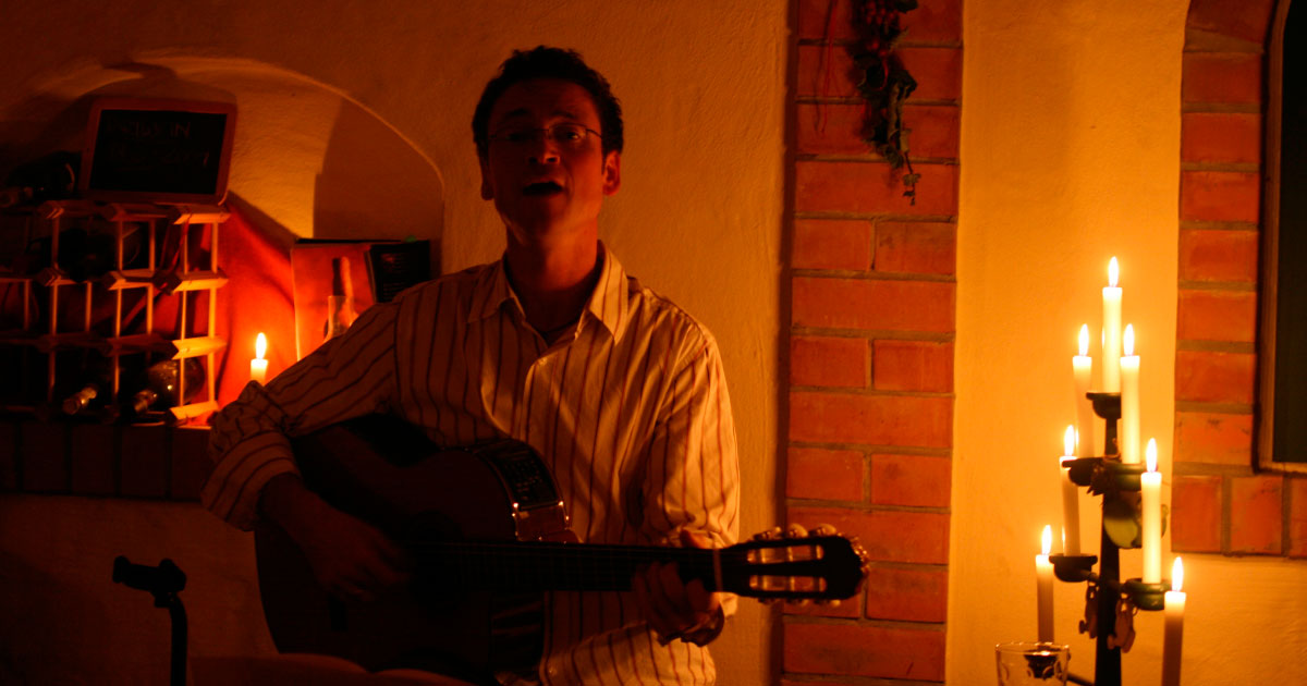 Mann sitzt auf einem Hocker und spielt Gitarre in gemütlicher Atmosphäre. Neben ihm brennt ein Kerzenleuchter. 