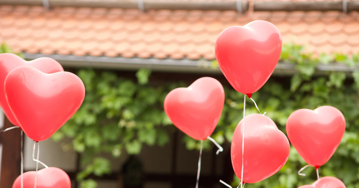 Im Vordergrund fliegen rosa Herz-Ballons mit einer weißen Schnur nach oben. Im Hintergrund sieht man ein rotes Dach und grüne Weinblätter an einem Gebäude 